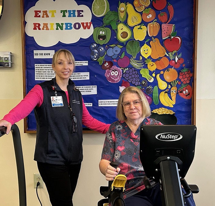 A female exercise physiologist stands next to a female cardiac rehab patient on an arm bike.