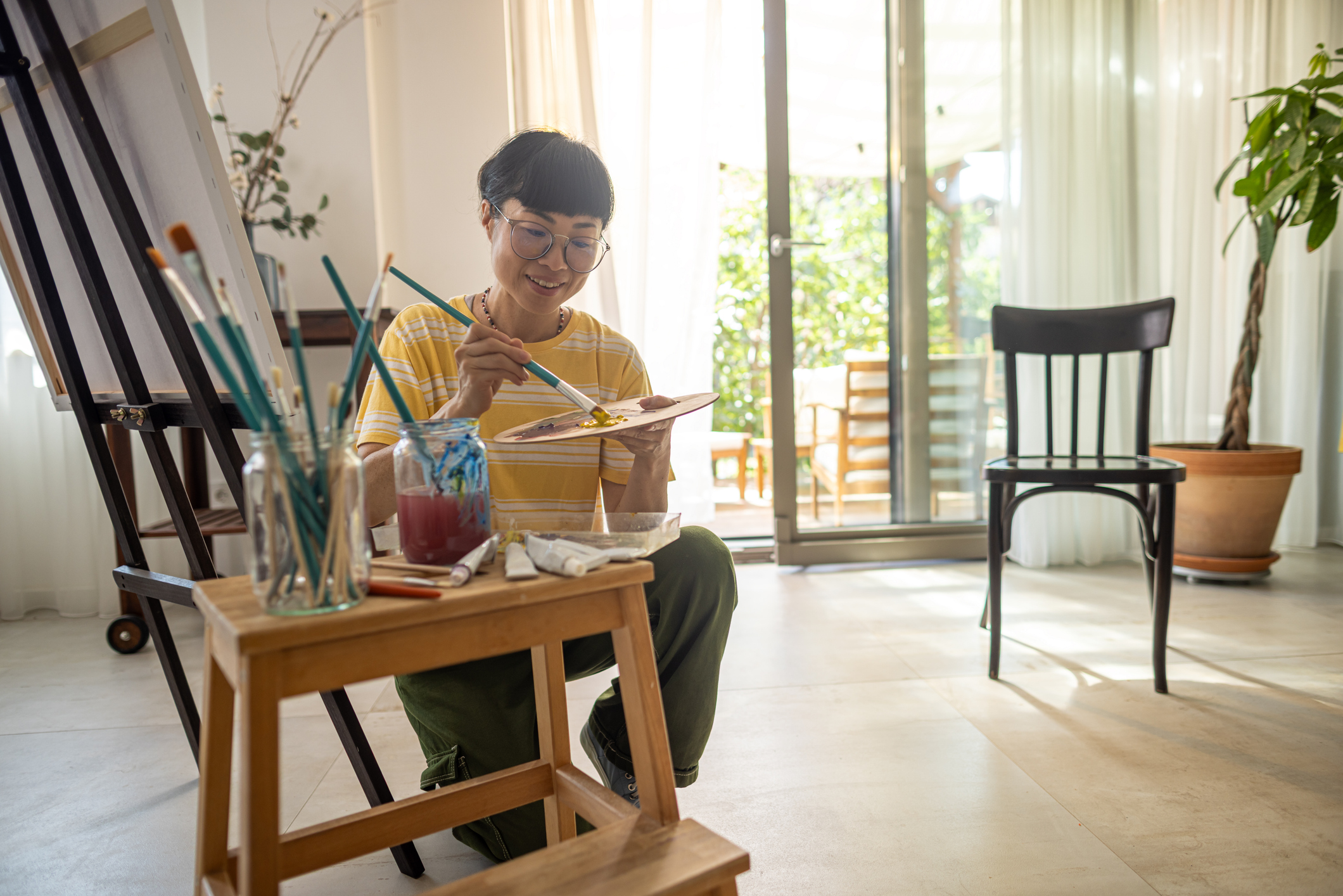 An older woman kneels down in her living room, smiling as she paints on a canvas