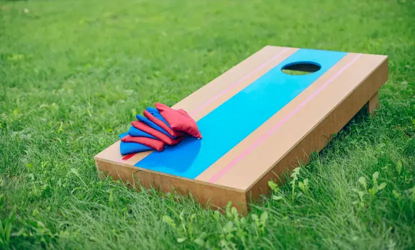 A red and blue wooden cornhole board sits on a green lawn, with eight alternating blue and red cornhole bags sitting in the lower corner.