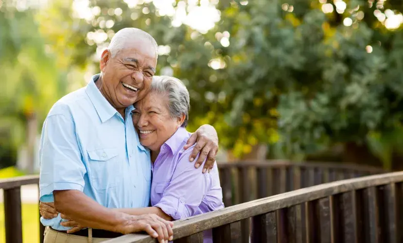 An older couple standing on a wooden bridge in nature laughs as they hug one another