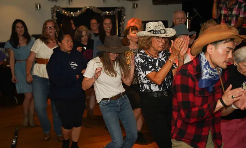 A mixed group of people in country attire line dancing at Kickin' Up Country 2025