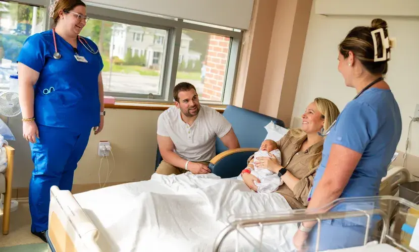 Two nurses look on as a new mom holds her infant in a hospital bed at South Shore Hospital with her husband next to her.