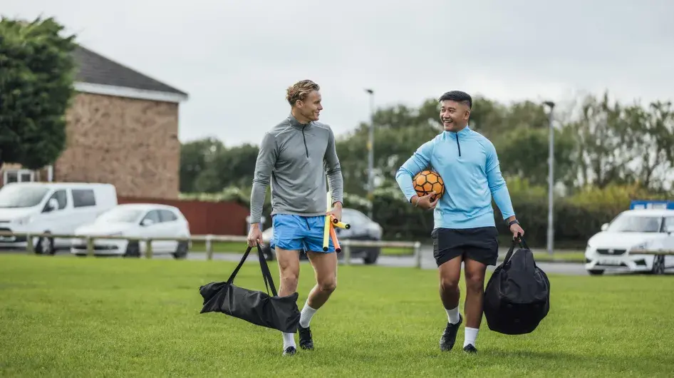 Two men smile as they talk to each other walking onto a soccer field, each carrying a bag, with balls, corner flags, etc.