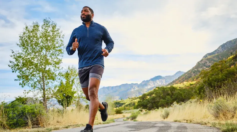 A middle-aged Black man looks happy as he jogs along an asphalt path in a hilly/mountain looking setting