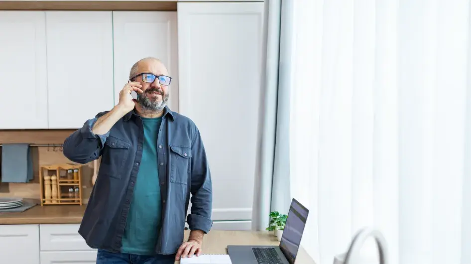 An older man stands at an open laptop in his kitchen while talking on the phone and smiling
