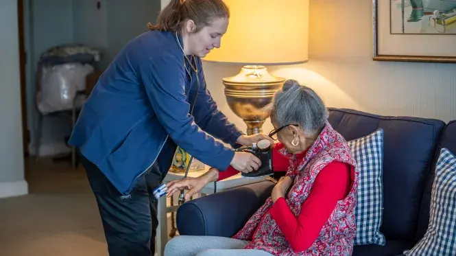 An older woman sits on her couch as a caregiver from South Shore VNA adjusts a blood pressure cuff on her right arm.