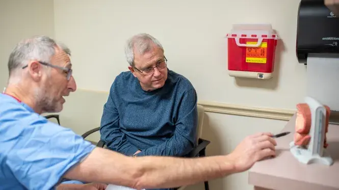 Sergio Fefer, MD points to an anatomical model as he speaks to an older patient seated in a chair.
