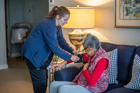An older woman sits on her couch as a caregiver from South Shore VNA adjusts a blood pressure cuff on her right arm.