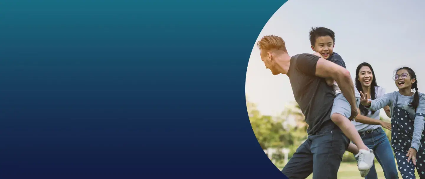 A dad carries his young son on his back as the mom and daughter chase them while playing tag.