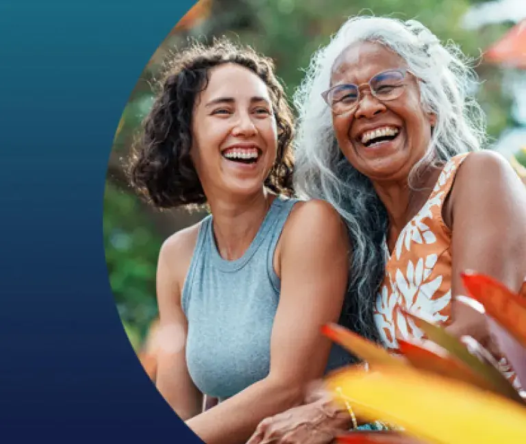 A mom and her adult daughter laugh as they sit next to one another in an outdoor setting.
