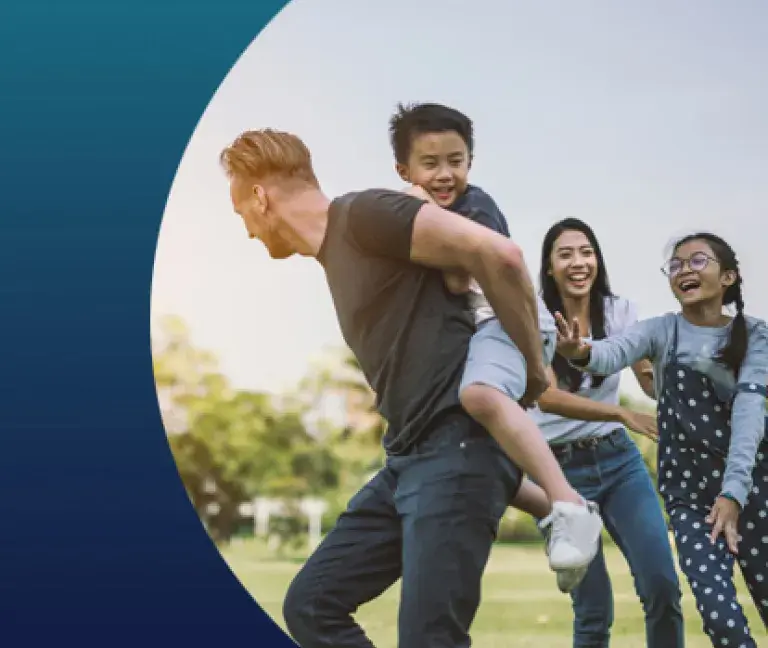 A dad carries his young son on his back as the mom and daughter chase them while playing tag.
