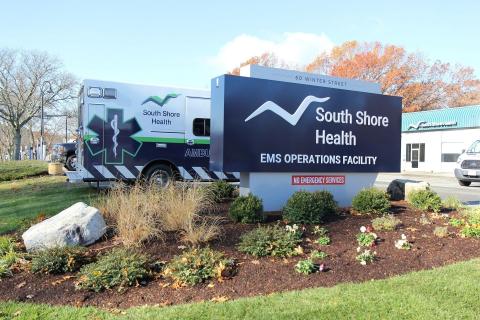 A sign at the exterior of South Shore Health EMS' Operations Facility at 60 Winter Street in Weymouth. The sign features the South Shore Health logo and text reading "EMS Operations Facility." An ambulance can be seen to the left.