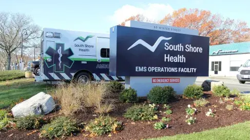 A sign at the exterior of South Shore Health EMS' Operations Facility at 60 Winter Street in Weymouth. The sign features the South Shore Health logo and text reading "EMS Operations Facility." An ambulance can be seen to the left.