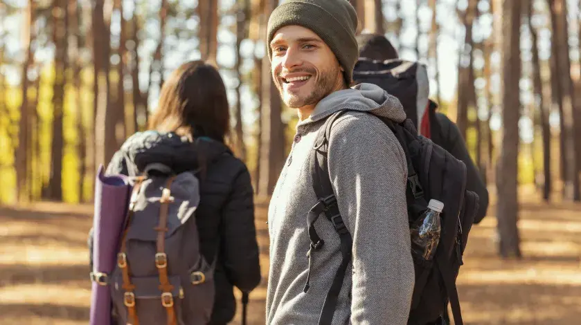 A man wearing fall-warm clothes smiles at the camera during a hike in the woods as two friends walk behind him
