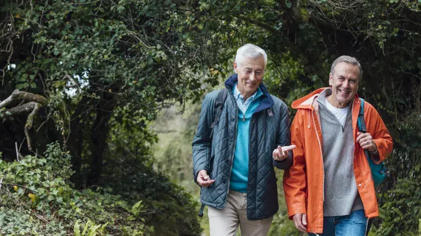 Two older men smile as they walk together on a hike through a forested area