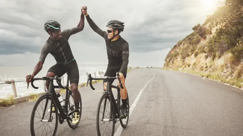 Two men smile as the high five while cycling down a coastal trail with the sun peeking out in the background