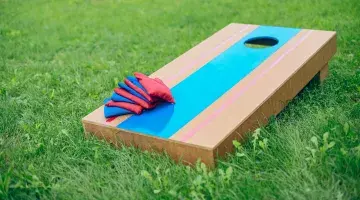 A red and blue wooden cornhole board sits on a green lawn, with eight alternating blue and red cornhole bags sitting in the lower corner.