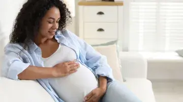 A pregnant woman sits on a couch gently cradling her belly in an image representing fetal movement monitoring, or Count the Kicks.
