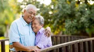 An older couple standing on a wooden bridge in nature laughs as they hug one another