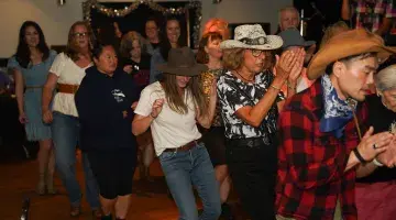 A mixed group of people in country attire line dancing at Kickin' Up Country 2025