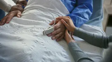 Close up view of a man's hands as he lies in a hospital bed and two people on either side of him gently place their hands on his hand and arm.