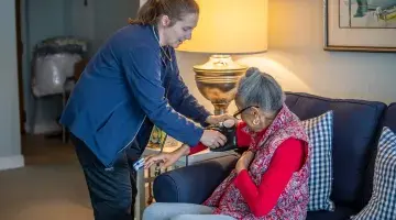 An older woman sits on her couch as a caregiver from South Shore VNA adjusts a blood pressure cuff on her right arm.