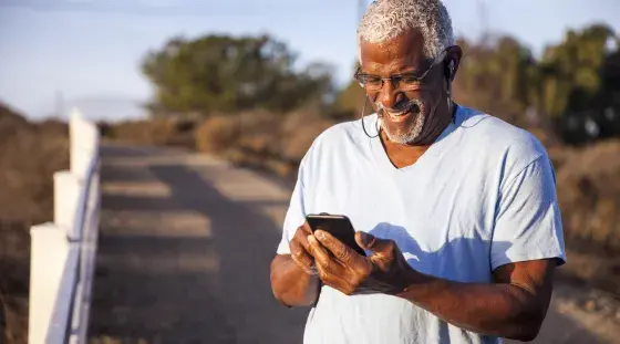 An older man out on a walk stops and smiles as he looks at the cell phone in his hand.