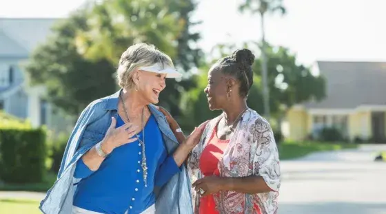 Two older women smile as they talk while walking down the street in an outdoor retirement community setting.
