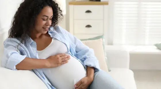 A pregnant woman sits on a couch gently cradling her belly in an image representing fetal movement monitoring, or Count the Kicks.
