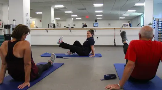 Jen lifts her leg to demonstrate a leg stretch as two participants look on during a community exercise class at South Shore Health